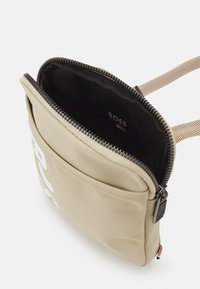 Borsa a tracolla in tessuto beige con zip nera, tasca frontale, tracolla regolabile e logo "BOSS" all'interno e sul cursore della zip.