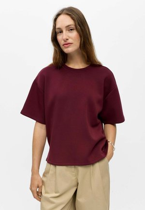 Femme aux longs cheveux bruns portant un haut bordeaux ample à manches courtes et un pantalon beige, debout, une main dans la poche, sur fond blanc.