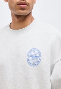 Νέος άνδρας που φοράει ανοιχτό γκρι φούτερ με μπλε κυκλικό λογότυπο "Bjorn Borg Stockholm Racquet and Health Club" στο στήθος.