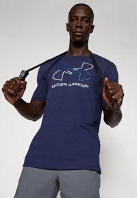 Navy blue cotton t-shirt featuring a large white "Under Armour" logo. The model holds resistance bands, wearing gray athletic shorts.