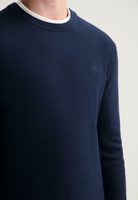 Marineblauer Strickpullover mit texturiertem Finish, rundem Halsausschnitt und dezentem Logo-Detail. Lange Ärmel und gerippte Bündchen runden das Design ab.