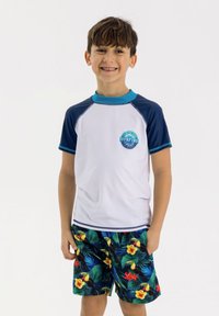 Pojkarnas simoutfit med en vit rash guard med kort ärm som har en blå logotyp, tillsammans med färgglada badshorts med tropiskt tryck.