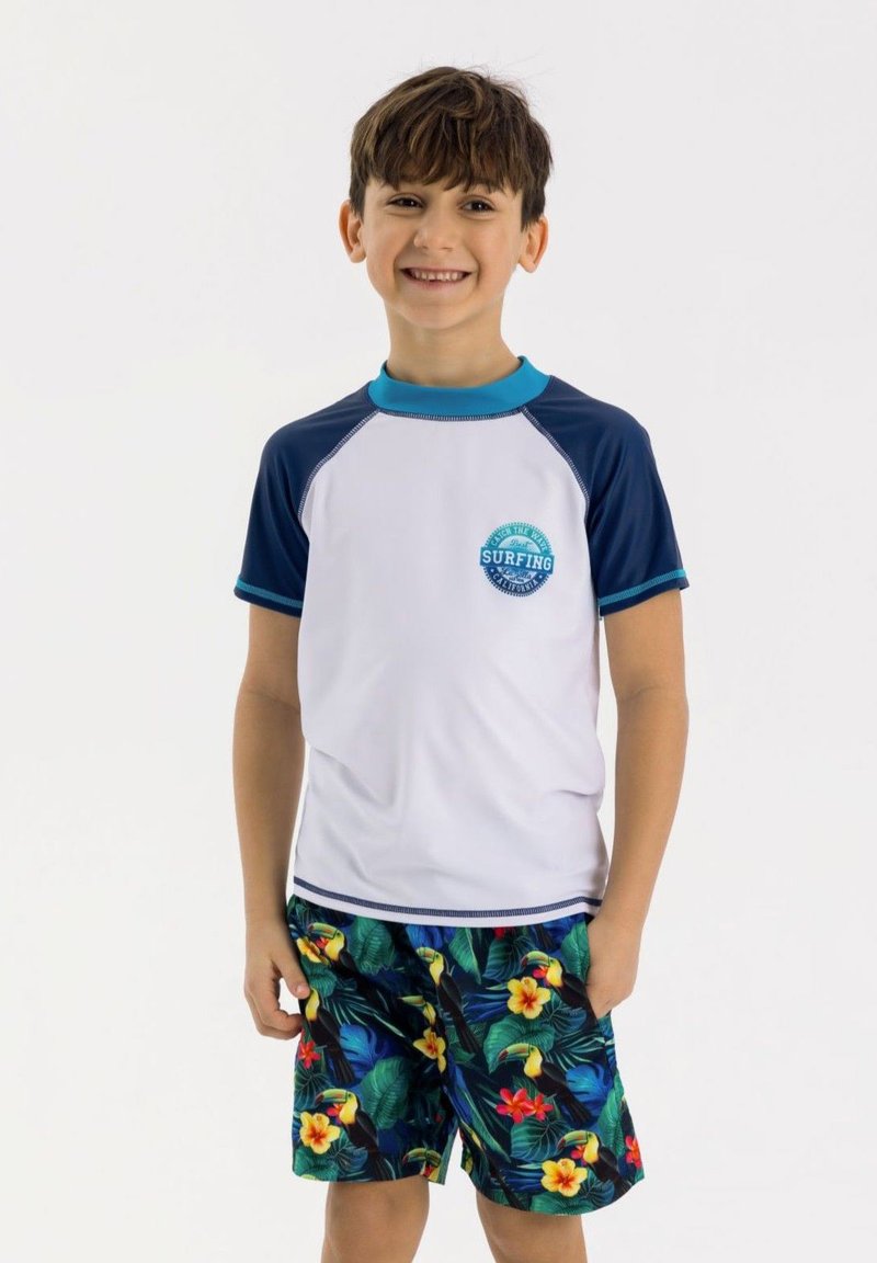 Pojkarnas simoutfit med en vit rash guard med kort ärm som har en blå logotyp, tillsammans med färgglada badshorts med tropiskt tryck.