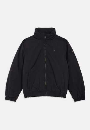 Črna jakna z zadrgo, stojnim ovratnikom, elastičnimi manšetami in spodnjim robom, sprednjimi žepi ter majhno rdeče-belo-modro oznako na prsih in levem rokavu.