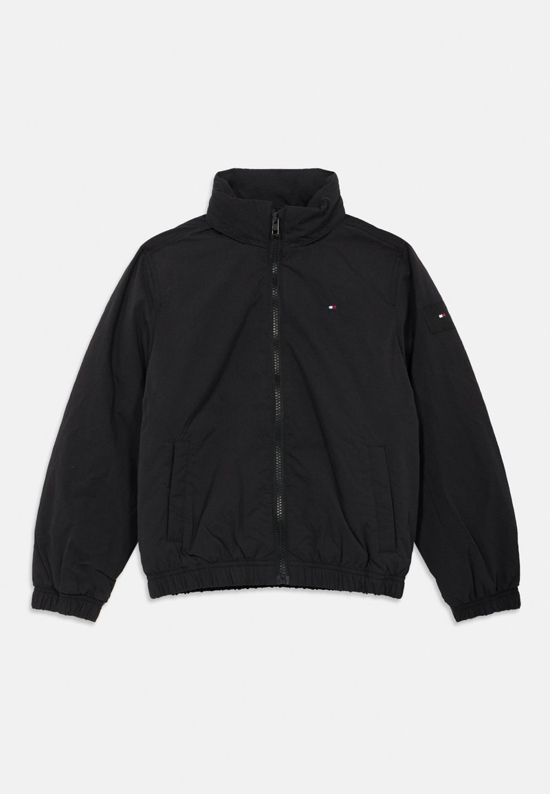 Črna jakna z zadrgo, stojnim ovratnikom, elastičnimi manšetami in spodnjim robom, sprednjimi žepi ter majhno rdeče-belo-modro oznako na prsih in levem rokavu.