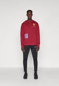 Κρασί φούτερ με στρογγυλή λαιμόκοψη, χρυσό έμβλημα και μπλε γραφικό πATCH. Συνδυασμένο με μαύρες φόρμες και μαύρα αθλητικά παπούτσια.