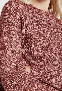 In primo piano, una persona indossa un spesso maglione a maglia color bordeaux e crema con le braccia incrociate, mostrando il motivo del tessuto testurizzato.