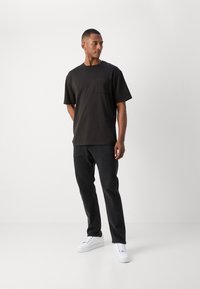 Mand iført en sort t-shirt med korte ærmer og brystlomme, mørke jeans og hvide sneakers, stående mod en ensfarvet lys baggrund.