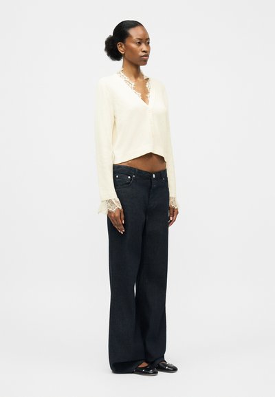 Γυναίκα που φοράει κρεμ πουλόβερ με δαντελωτό V-λαιμόκοψη, φαρδιά μαύρα τζιν και μαύρα παπούτσια χωρίς κορδόνια μπροστά σε απλό φόντο.