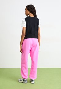 Svart strikket cropped vest over en hvit t-skjorte, kombinert med sterke rosa joggebukser og grå sneakers, med en jevn tekstur og avslappet passform.