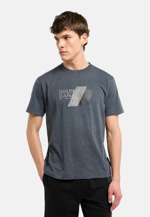 Ung mand iført en mørkegrå Bruno Banani T-shirt med et geometrisk logo og sorte bukser, stående mod en ensfarvet hvid baggrund.