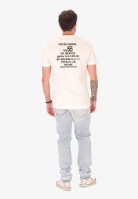 Uomo con braccio destro tatuato che indossa una t-shirt bianca stampata, jeans azzurri chiari e sneakers bianche, visto da dietro su uno sfondo neutro.