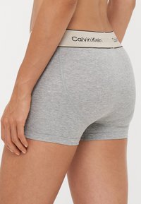 Sive pamukaste bokserice s teksturiranim završetkom, koje imaju pojas u crnoj i krem boji s logotipom Calvin Klein.