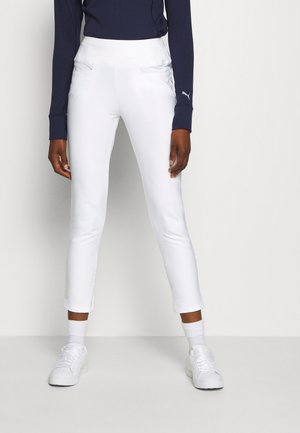 Osoba ubrana w dopasowane białe spodnie, białe trampki oraz granatową bluzkę z długim rękawem, stojąca na tle jasnego, jednolitego tła.