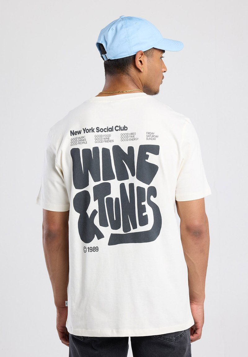 Crème-kleurige katoenen t-shirt met gedurfde zwarte tekst op de achterkant: "WINE & TUNES" met aanvullende kleinere tekst erboven en eronder.