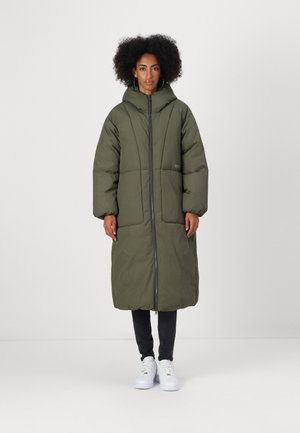 Manteau matelassé vert olive avec une capuche, manches longues et fermeture éclair avant. Il présente un design matelassé et des poignets côtelés, porté sur un pantalon sombre et des chaussures blanches.
