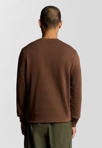 Barna hosszú ujjú pulóver, kerek nyakkivágással és bordázott mandzsettákkal, zöld rakott nadrággal párosítva. Simább pamut anyag, minimális díszítéssel.