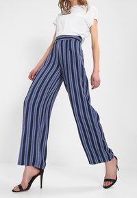 Pantalones de pierna ancha de color azul marino con rayas verticales en blanco y púrpura. Presentan una cintura alta y una textura de tela suave, combinados con tacones negros.
