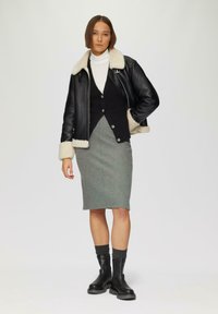 Veste en cuir noire avec col en shearling, pull à col roulé blanc, cardigan noir, jupe crayon grise et bottines noires avec des chaussettes côtelées.