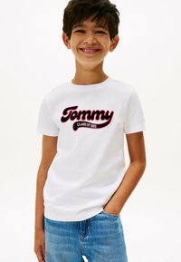T-shirt in cotone bianco con grafica "Tommy" e "CLASS OF 1985" in rosso e nero. Maniche corte e scollo rotondo, abbinato a jeans blu.
