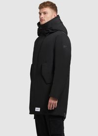 Cămașă parka neagră cu guler înalt, buzunare frontale și o pată de logo pe manșetă. Are o textură căptușită și detalii cu fermoar.