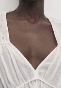 Chemisier blanc avec un décolleté en V profond, tissu doux et léger avec une texture subtile, et des détails froncés près du décolleté.