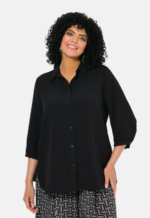 Camicia nera con bottoni, colletto, maniche a tre quarti e tessuto testurizzato. Abbinata a pantaloni a righe bianchi e neri.