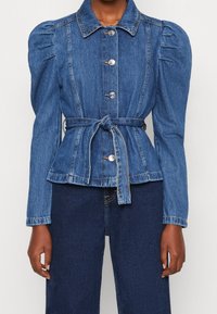 Giacca di jeans con maniche a sbuffo, chiusura con bottoni e cintura da annodare in vita. Colore azzurro chiaro con una finitura leggermente testurizzata e dettagli di cucitura visibili.