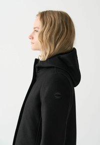 Juodas gobtuvas paltas, pagamintas iš tekstūruoto audinio, turintis apvalų logotipo ženklelį ant rankovės ir ploną siluetą.