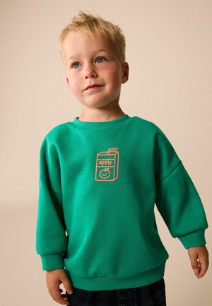Young boy with light hair wearing a green sweatshirt with an orange apple juice box graphic and black pants standing against a beige background.