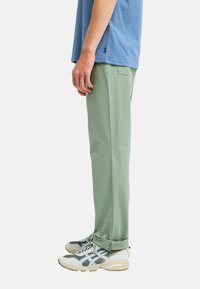 Man standing in green pants, blue t-shirt, and gray athletic sneakers, shown from side against white background.