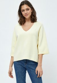 Maglione giallo, largo e morbido, con scollatura a V e maniche 3/4. Realizzato in tessuto testurizzato, presenta un orlo dritto. Abbinato a jeans blu.