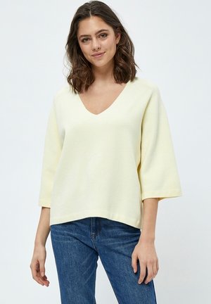 Maglione giallo, largo e morbido, con scollatura a V e maniche 3/4. Realizzato in tessuto testurizzato, presenta un orlo dritto. Abbinato a jeans blu.