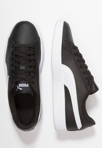 Baskets Puma noires avec une bande blanche et une semelle blanche, lacets noirs, et logo sur la languette, présentées vues de dessus et de côté sur fond blanc.