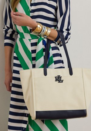 Žena v modro-bielych pruhovaných šatách so zelenými diagonálnymi pásmi, drží béžovú plátenú tašku s čiernymi rúčkami a logom RL.