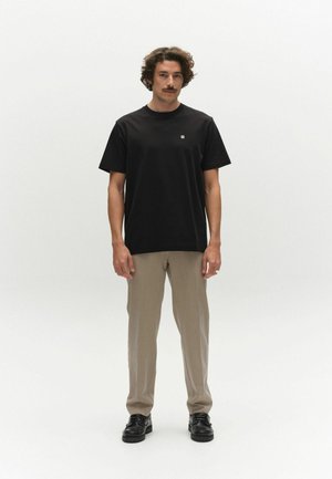 Mann steht vor weißem Hintergrund, trägt ein schwarzes kurzärmliges Hemd, beige Hose und schwarze Schuhe, schaut nach vorne mit entspannter Haltung.