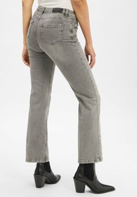 Graue Denim-Jeans mit schmaler Taille und ausgestellten Beinen. Enthält fünf Taschen und sichtbare Nähte. Kombiniert mit schwarzen, knöchelhohen Absatzstiefeln.