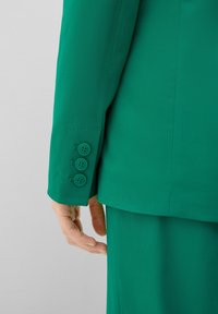 Green tailored suit jacket sleeve showing three green buttons and a clean seam finish. The fabric appears smooth with a matte texture.