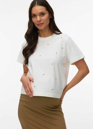 Woman with long dark hair wearing a white short-sleeve shirt with small yellow flower prints and a brown skirt, posing with one hand on her hip.