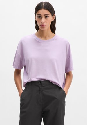 Woman with straight dark hair wearing a loose lavender short-sleeve shirt tucked into high-waisted black pants with hands in pockets.