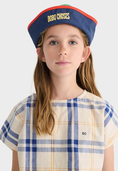 Jeune fille aux longs cheveux portant un chapeau de marin bleu bordé d'orange avec l'inscription "BOBO CHOSES" et une robe beige à motifs écossais bleu et orange.