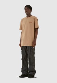 Lys beige t-skjorte med korte ermer og diskret logo, kombinert med løstsittende, mørke cargobukser med flere lommer og en teksturert overflate.
