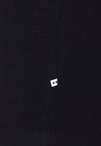 Tissu noir texturé avec un motif gaufré subtil ; présente une petite étiquette logo blanche dans le coin inférieur.