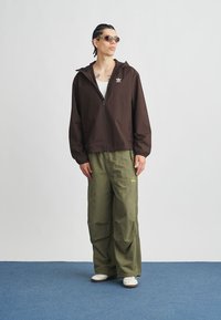 Καφέ ζακέτα με φερμουάρ και κουκούλα, με λευκό λογότυπο· φαρδιά χακί παντελόνια cargo· λευκά αθλητικά παπούτσια· καφέ γυαλιά ηλίου.