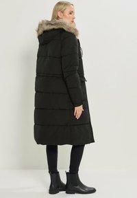 Abrigo acolchado negro con capucha de piel sintética desmontable, diseño acolchado horizontal, combinado con pantalones negros ajustados y botas de tobillo.