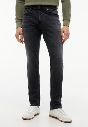 Homme portant un jean slim noir délavé, une chemise verte à manches longues et des baskets blanches avec semelles marron, debout sur un fond blanc.
