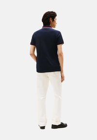 Camiseta tipo polo azul marino con detalles en rojo y blanco en el cuello y las mangas. Combinada con pantalones blancos y zapatos negros, de espaldas al espectador.