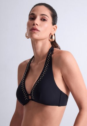 Femme portant un haut de bikini noir à col halter avec des clous dorés et de grandes boucles d'oreilles créoles dorées sur un fond clair uni.