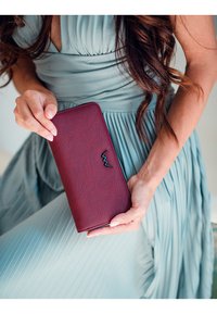 Burgundy leather wallet with a smooth, textured surface and a sleek black logo on the front, held against a pleated mint dress.