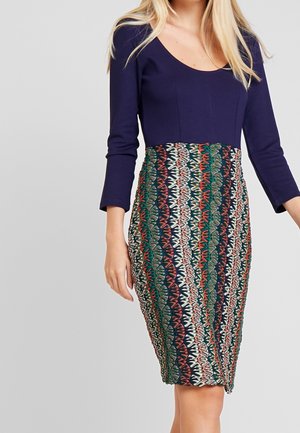 Långärmad klänning med en marinblå överdel och en figurnära, mönstrad kjol med geometriska mönster i gröna, röda och krämfärgade nyanser.
